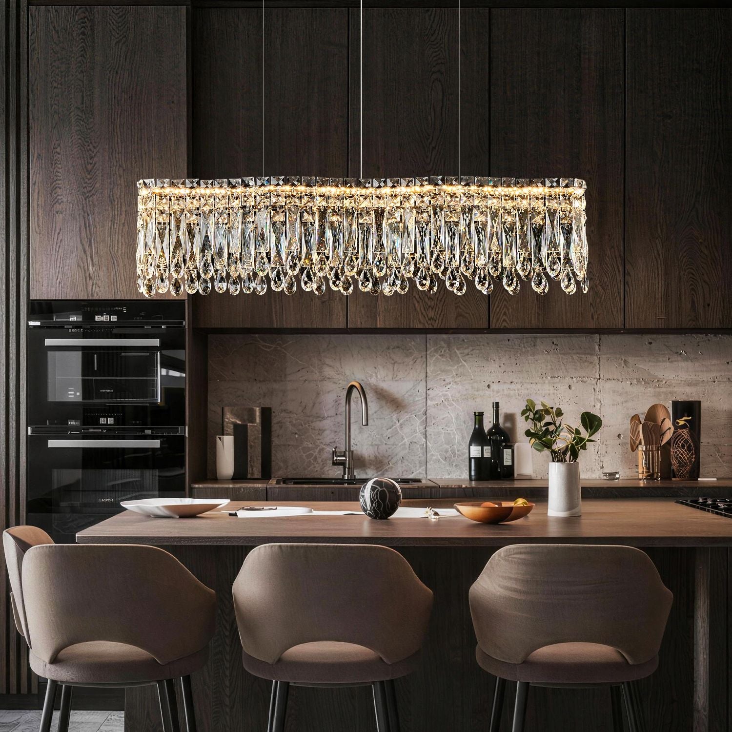 Flow Linear Modern Wave Integrated LED K9 Crystal Dining Chandelier in a kitchen island — hanging above a marble island with white flowers, elegant kitchenware, and dark wood cabinetry | Sofary Lighting