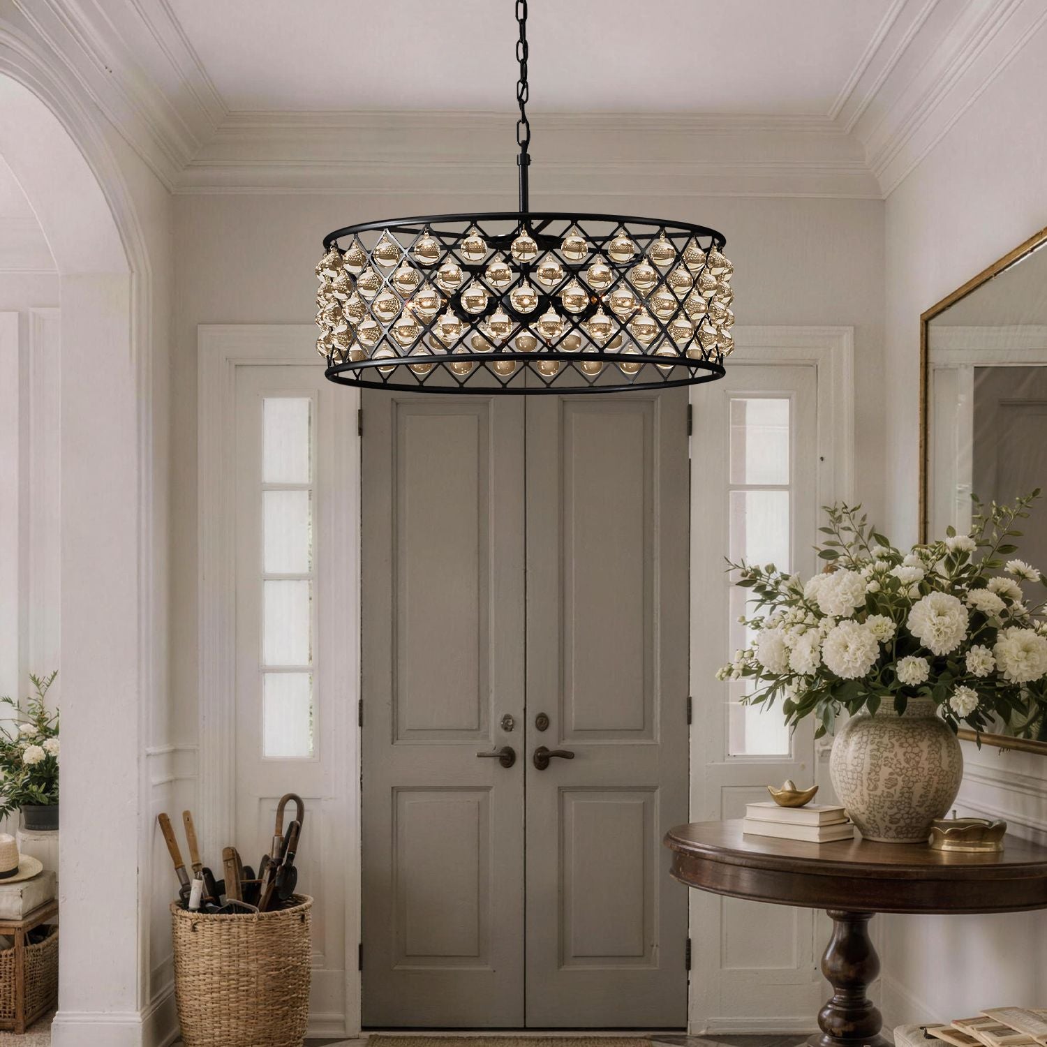 Harlow Vintage Matte Black K9 Crystal Round Chandelier in a foyer — suspended above a wooden console table with a vase of white flowers, books, and a gray double door backdrop | Sofary Lighting