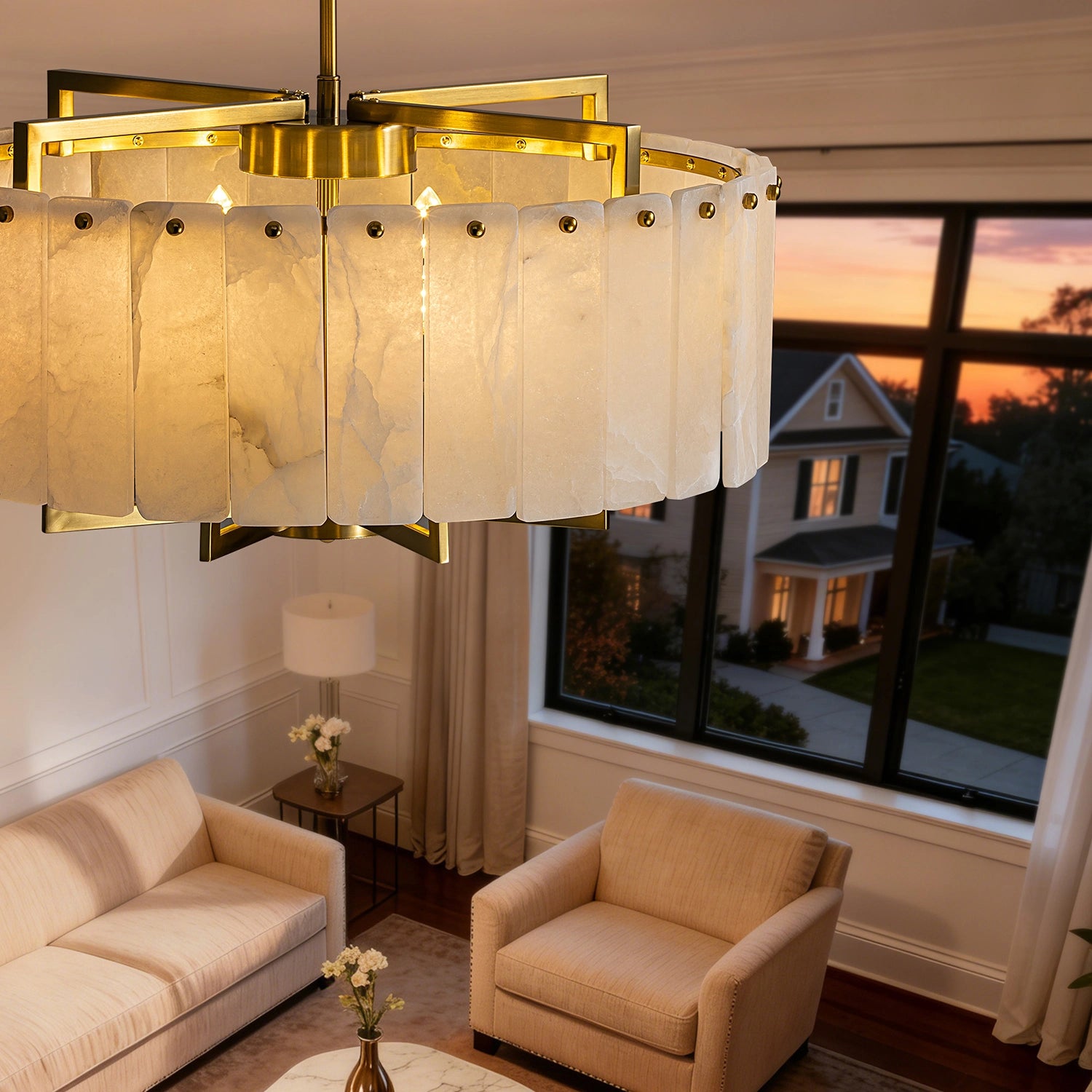 High-angle view of a round white alabaster chandelier in a luxury living room, showing the gold frame detail and soft ambient glow against a sunset window view. Sofary Lighting