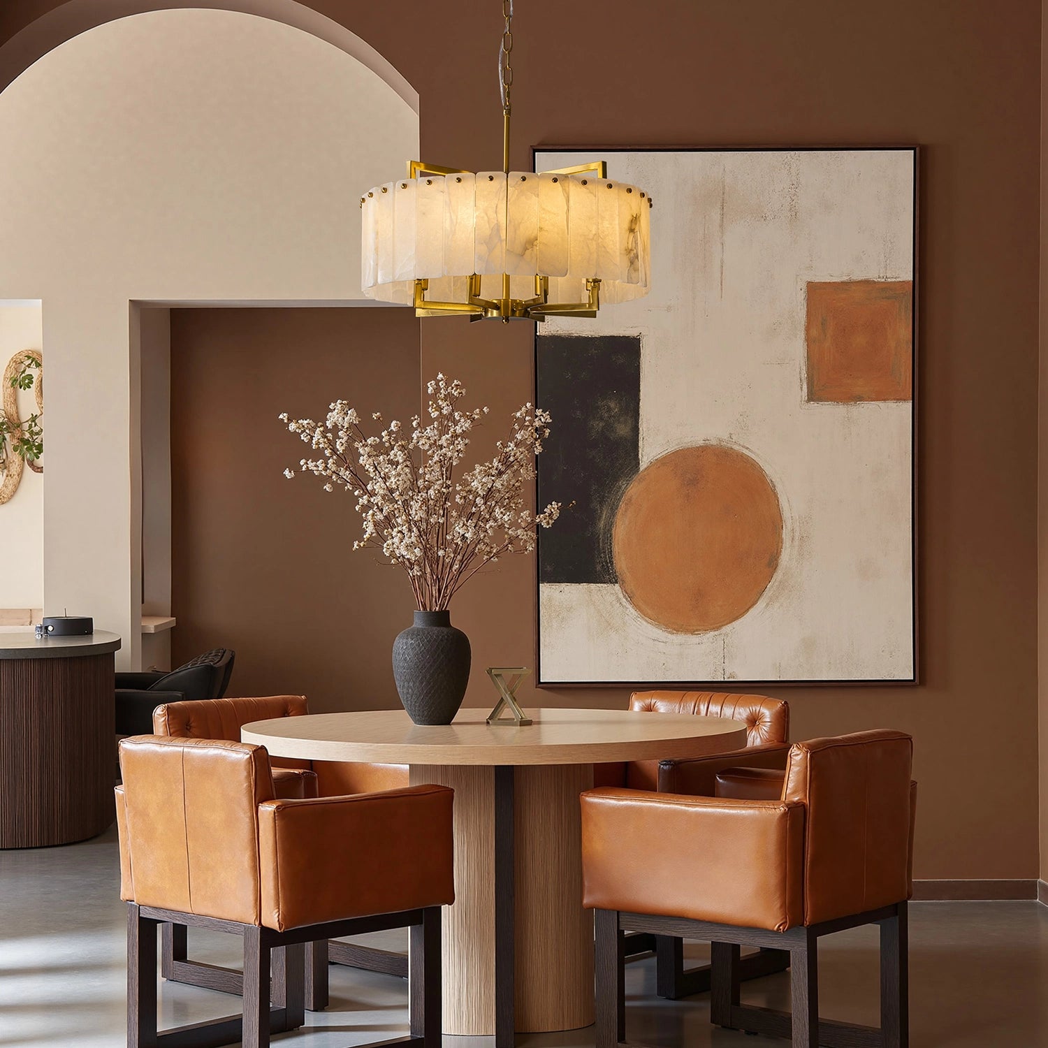 Circular alabaster stone chandelier with gold frame in a modern earthy dining room featuring terracotta walls, leather chairs, and minimalist abstract art. Sofary Lighting