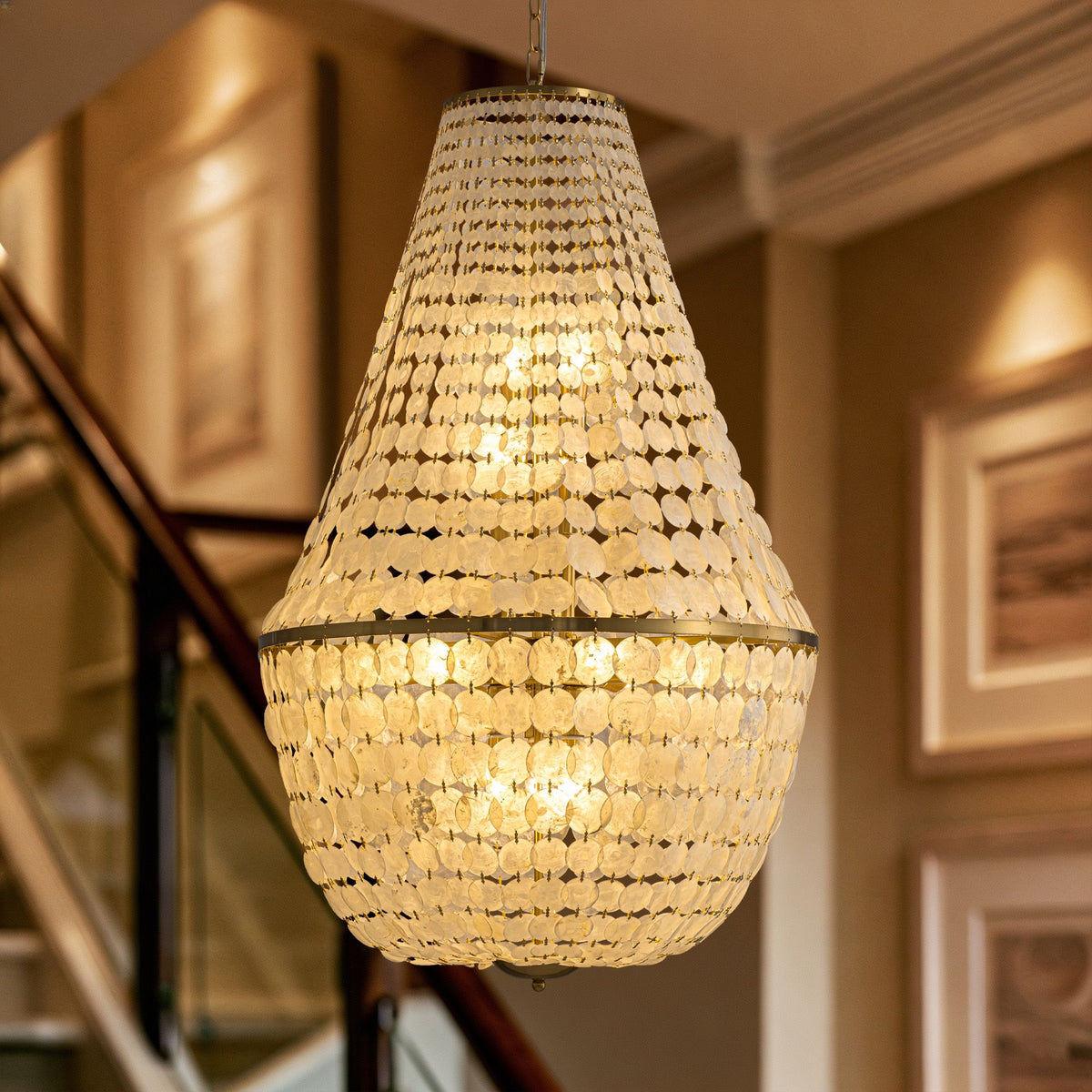 Aeroh Shell Coastal Gold Chandelier — teardrop/pear-shaped pendant, gold metal frame, cascading white circular shell/pearl discs, warm glowing light, staircase background, coastal luxury lighting | Sofary Lighting