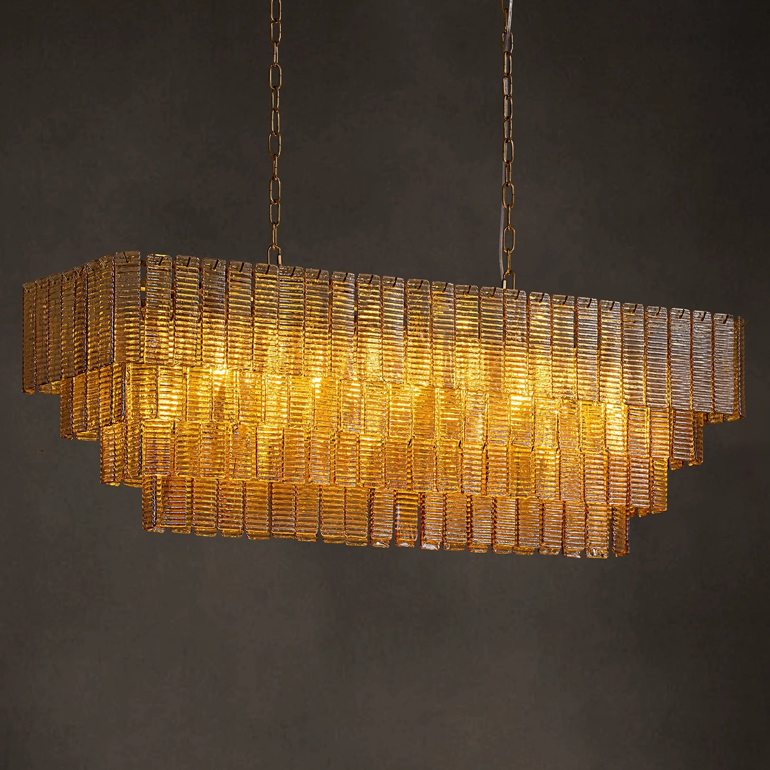 Rectangular tiered amber glass chandelier with a warm golden glow and brass hanging chains against a neutral grey background. Sofary Lighting