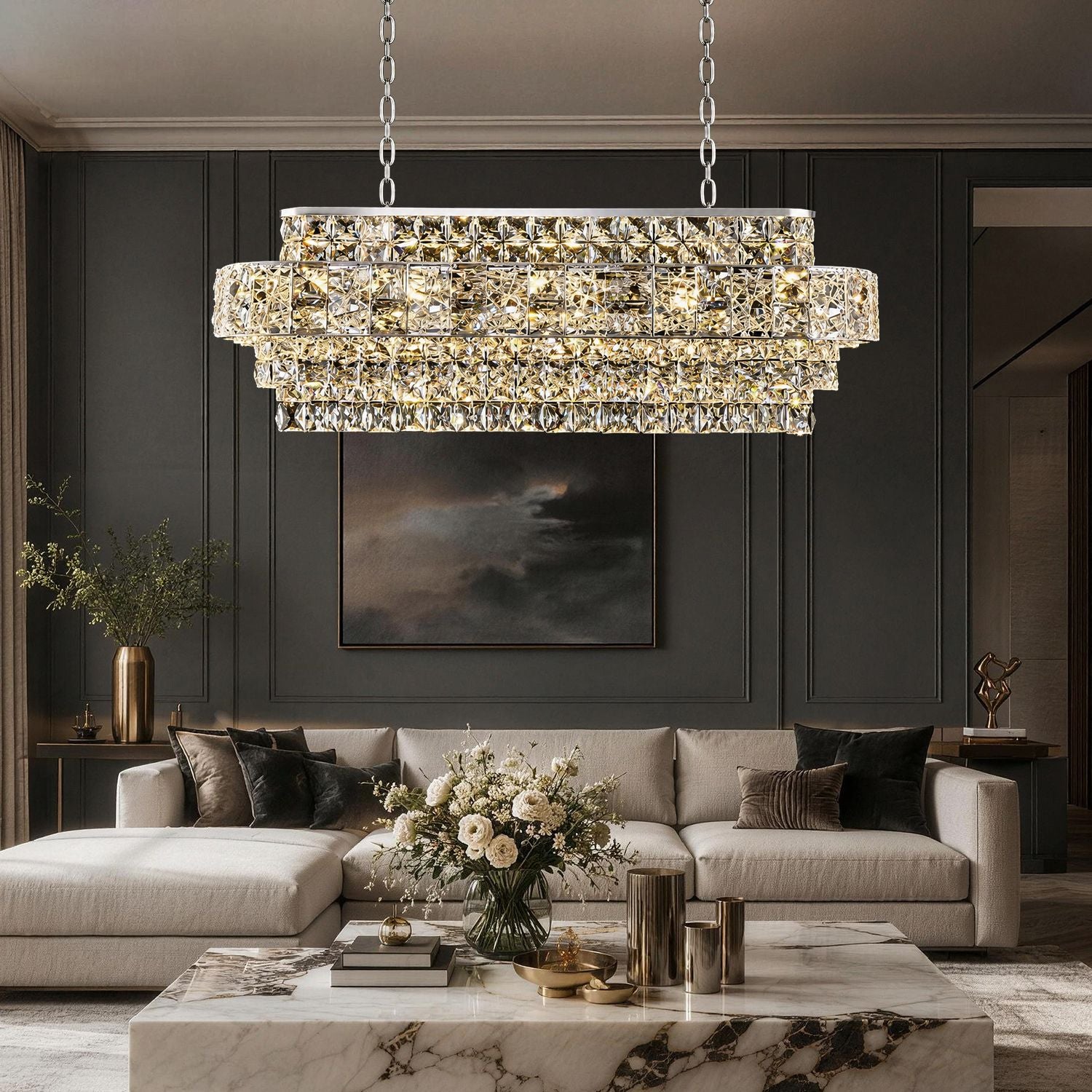 Valeria Linear Luxury K9 Crystal Chandelier in a living room — illuminating a beige L-shaped sofa with decorative pillows, a dark wood coffee table with books and sculptures